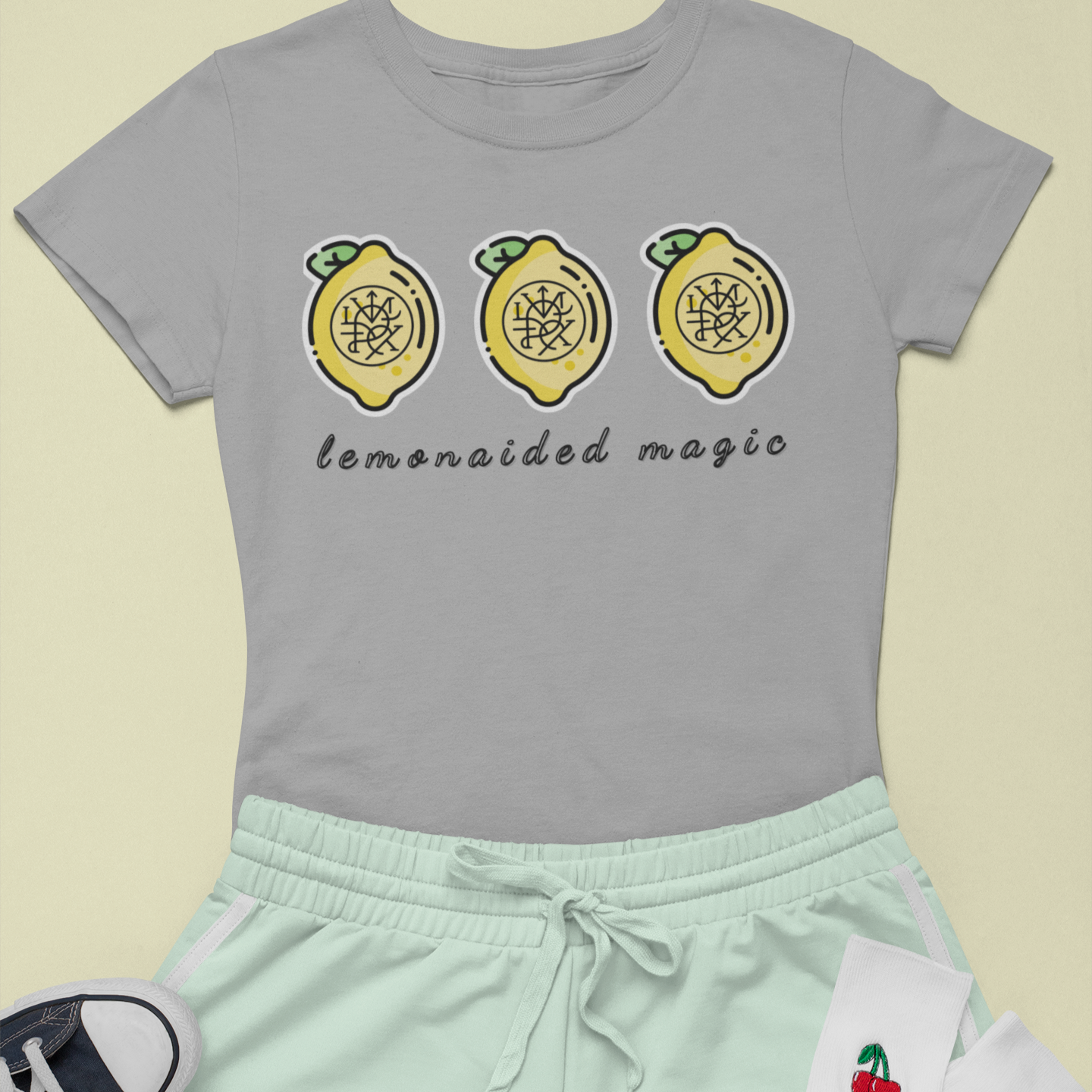 Flat lay of Lemonaided Magic custom sigil T-shirt — personalized affirmation shirt on lemon background Gray t-shirt with lemon design and text, mint shorts, white socks with cherry design, and black sneakers on a light yellow background.